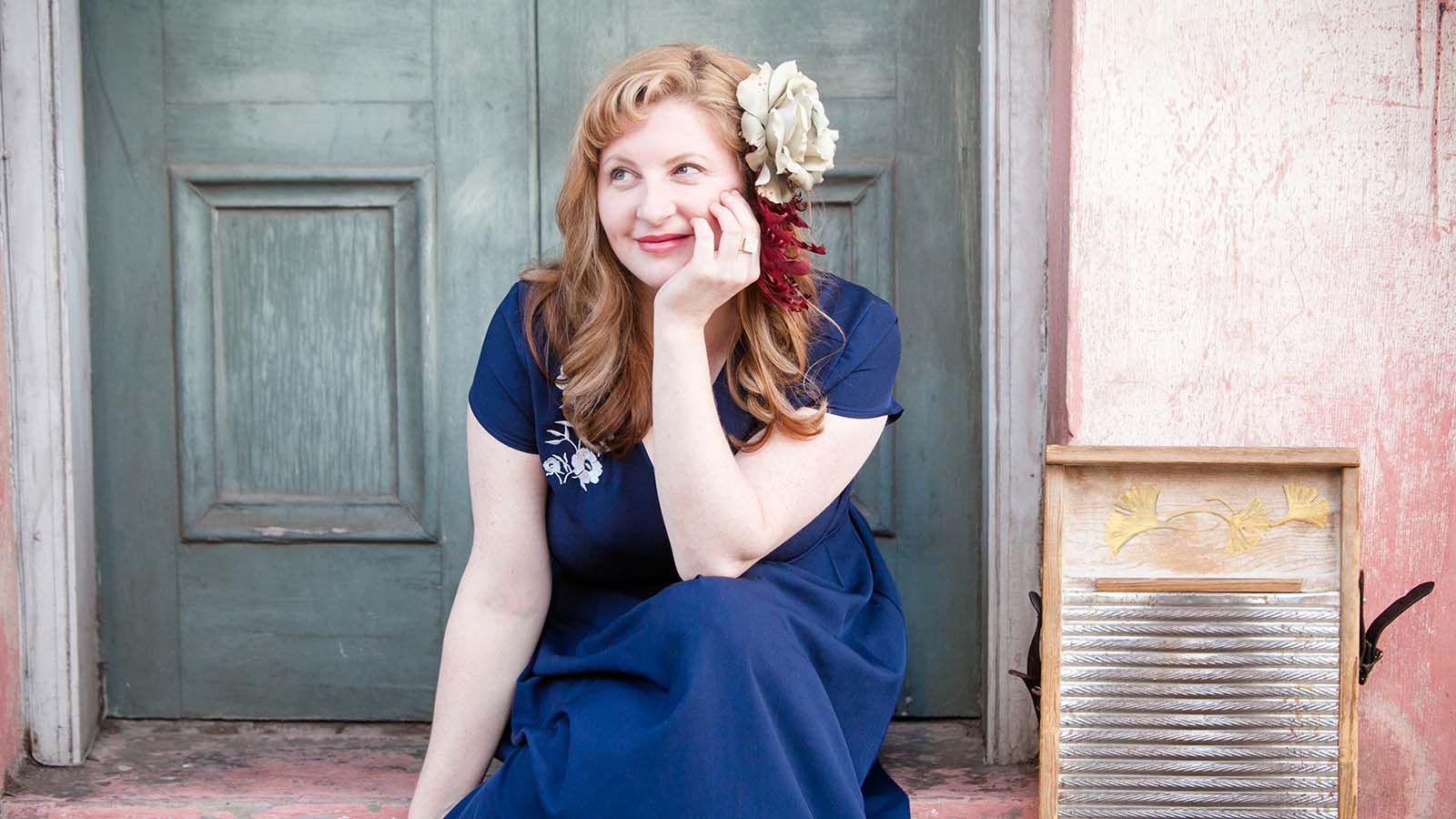 A woman with wavy hair sits on steps, wearing a navy dress adorned with floral embroidery, next to an old wooden washboard.