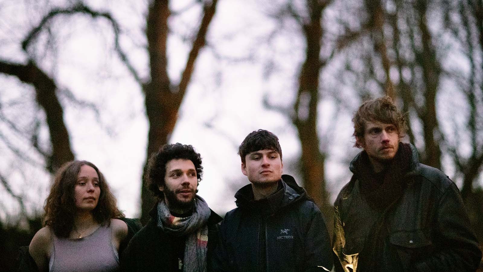 Four individuals stand together outdoors surrounded by trees at dusk, dressed in warm clothing, exuding a casual and intimate atmosphere.