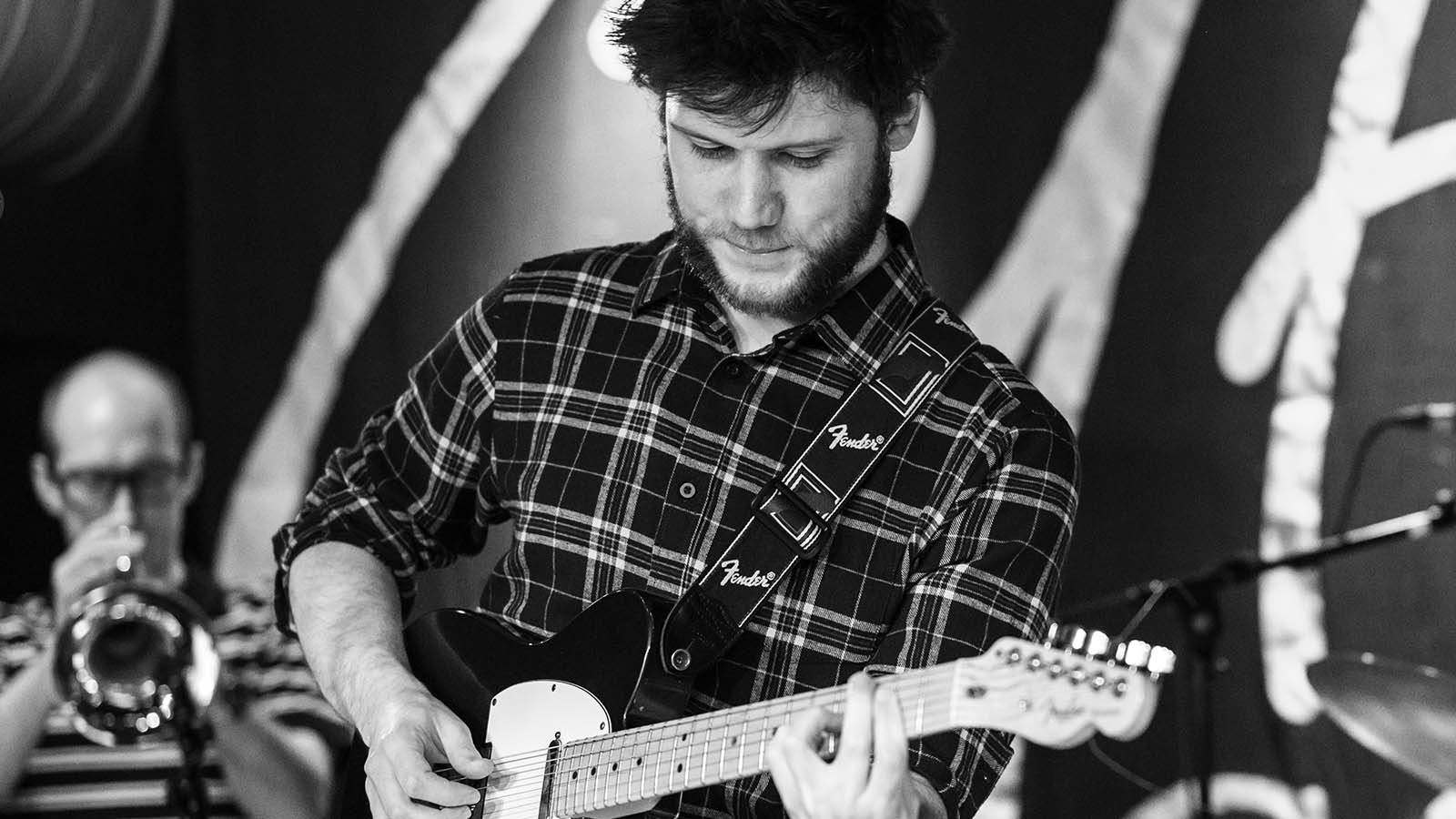A musician in a checkered shirt plays an electric guitar on stage, focusing intently as another performer plays a brass instrument behind him.