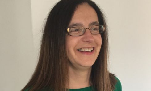 A woman with long brown hair wearing a green sweater is standing against a light-coloured wall.