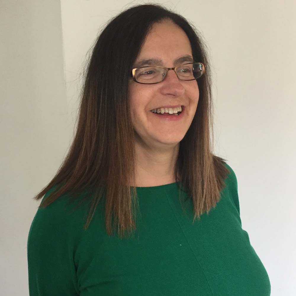 A woman with long brown hair wearing a green sweater is standing against a light-colored wall.