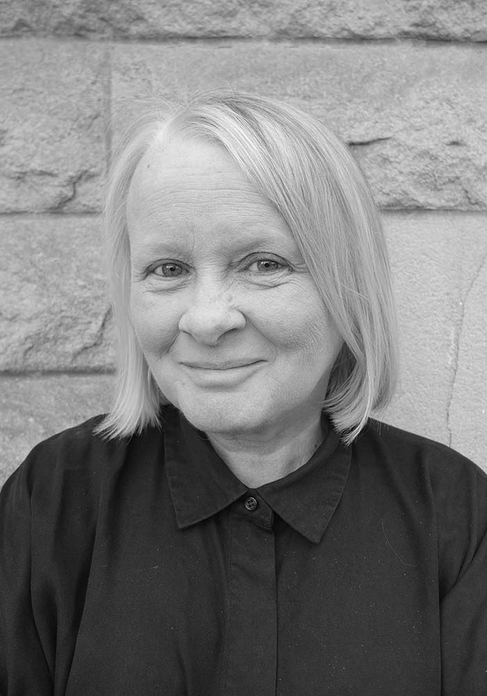 A woman with shoulder-length, gray hair wearing a dark collared shirt, stands in front of a textured stone wall.