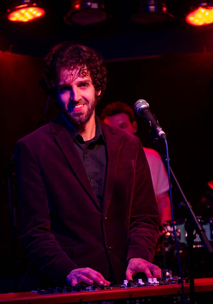 A performer stands on stage in a dimly lit venue, with a microphone in front of him, exuding an atmosphere of anticipation.