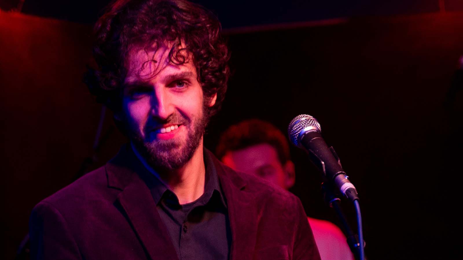 A performer stands on stage in a dimly lit venue, with a microphone in front of him, exuding an atmosphere of anticipation.