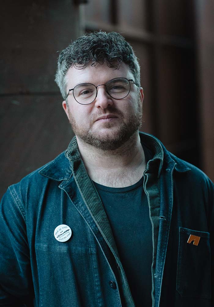 A person in a worn blue jacket and black t-shirt stands against a dark wooden backdrop, wearing a badge on their shirt.