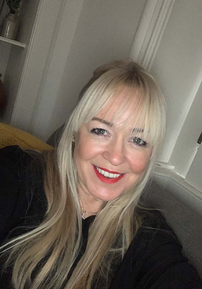 A person with long blonde hair is sitting on a couch, wearing a black top, with soft lighting illuminating the room.