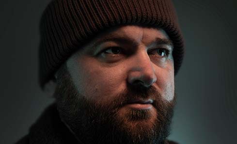A person wearing a dark knit hat and a textured coat stands in low light, creating a mysterious atmosphere.