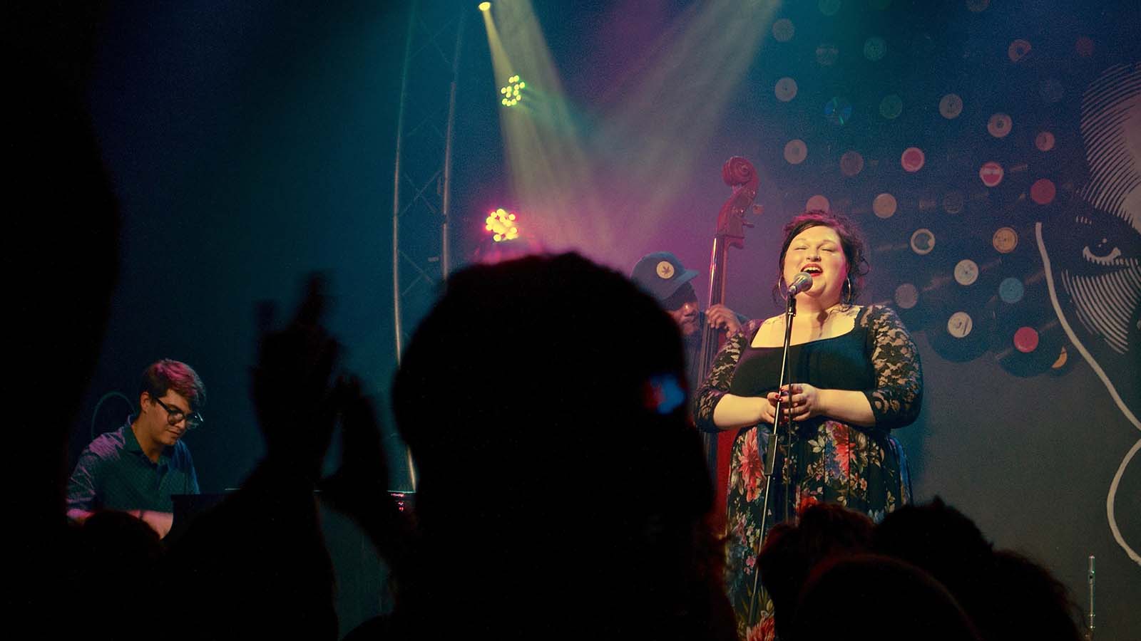 A singer performs on stage with a bassist, illuminated by colourful lights, while an excited audience raises their hands.
