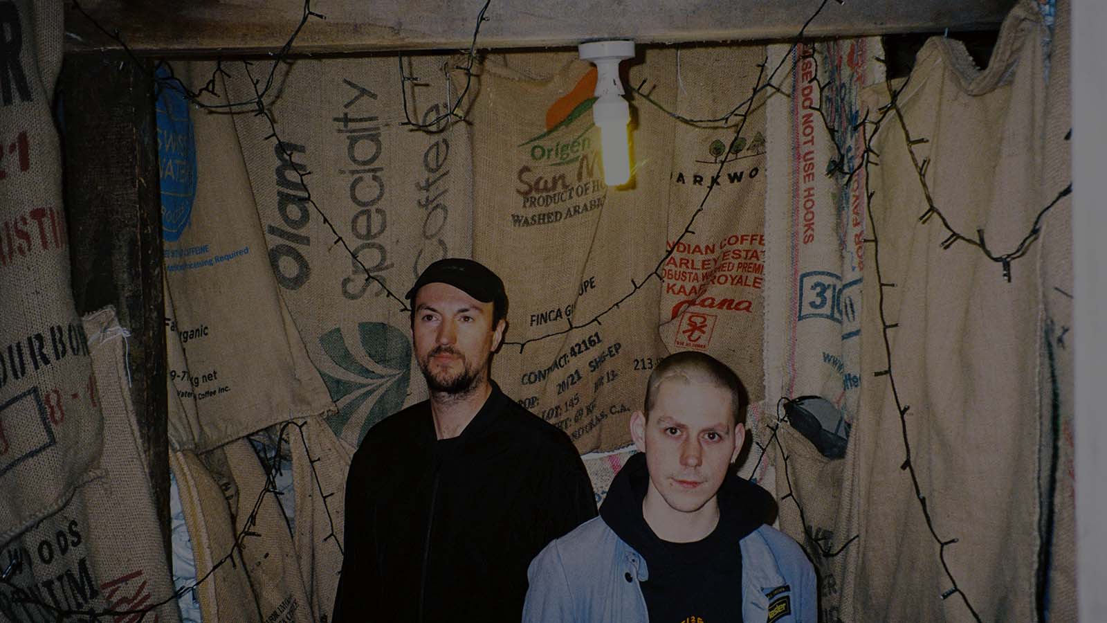 Two individuals stand inside a dimly lit room adorned with coffee sack walls and string lights, creating a cozy, rustic atmosphere.