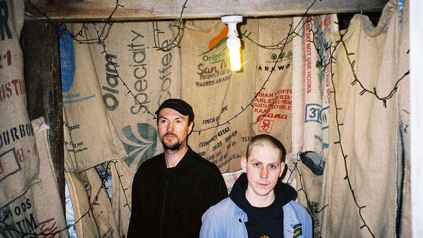 Two individuals stand inside a dimly lit room adorned with coffee sack walls and string lights, creating a cozy, rustic atmosphere.