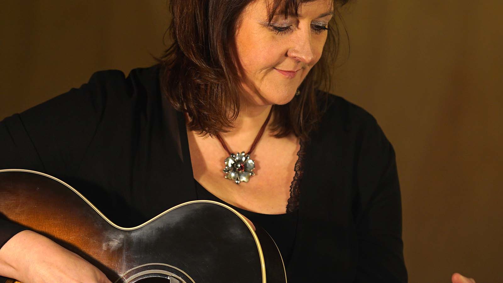 A woman in a black top holds a guitar, wearing a decorative necklace, with a blurred background that enhances the focus on her.