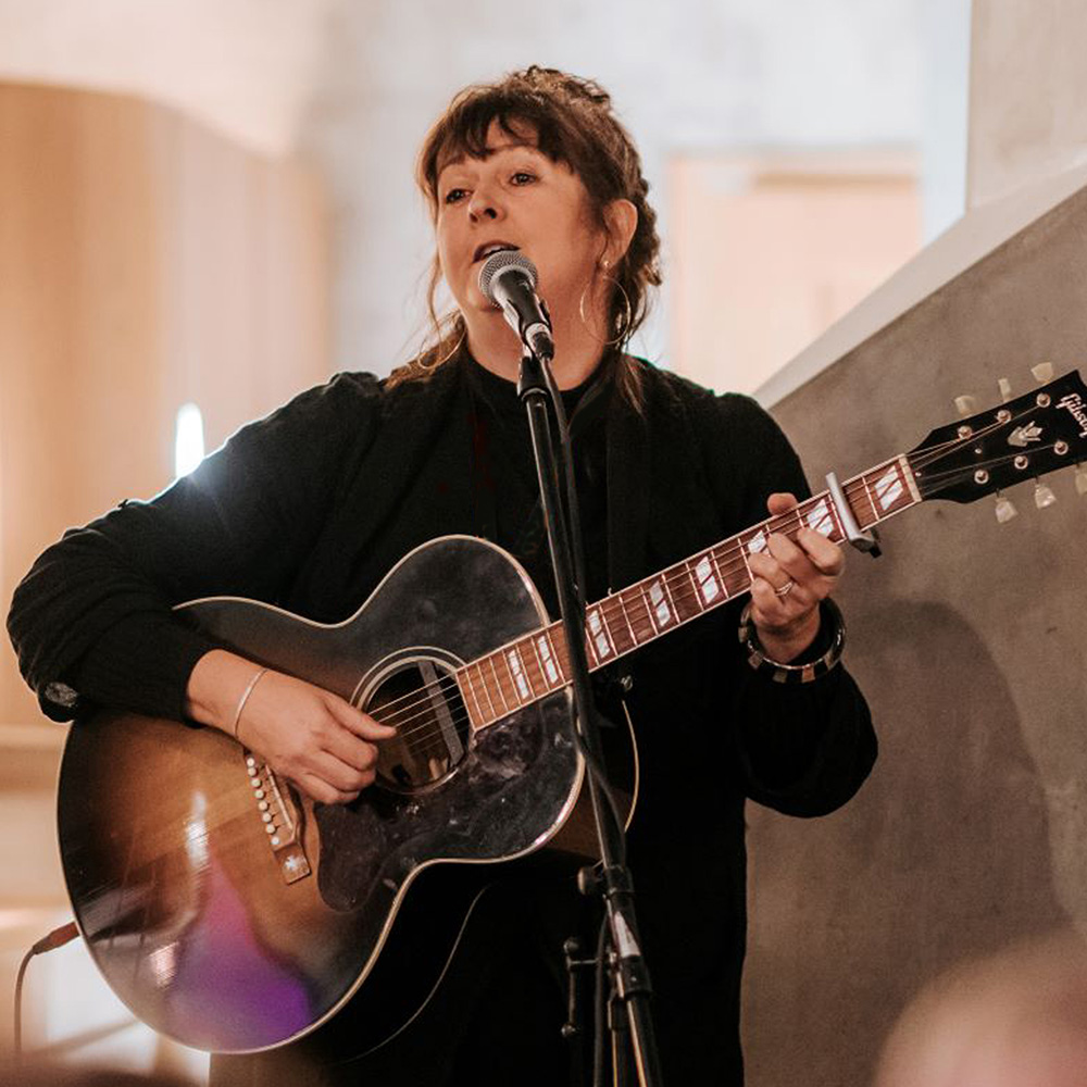 A performer sings and plays a rustic acoustic guitar on stage, with warm indoor lighting and modern decor in the background.