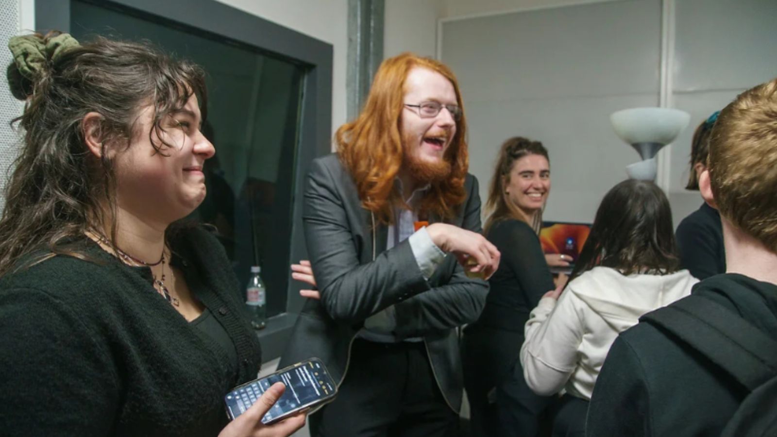 A group of people socialize in a modern, casual setting, with various individuals engaged in conversation and one person holding a phone.