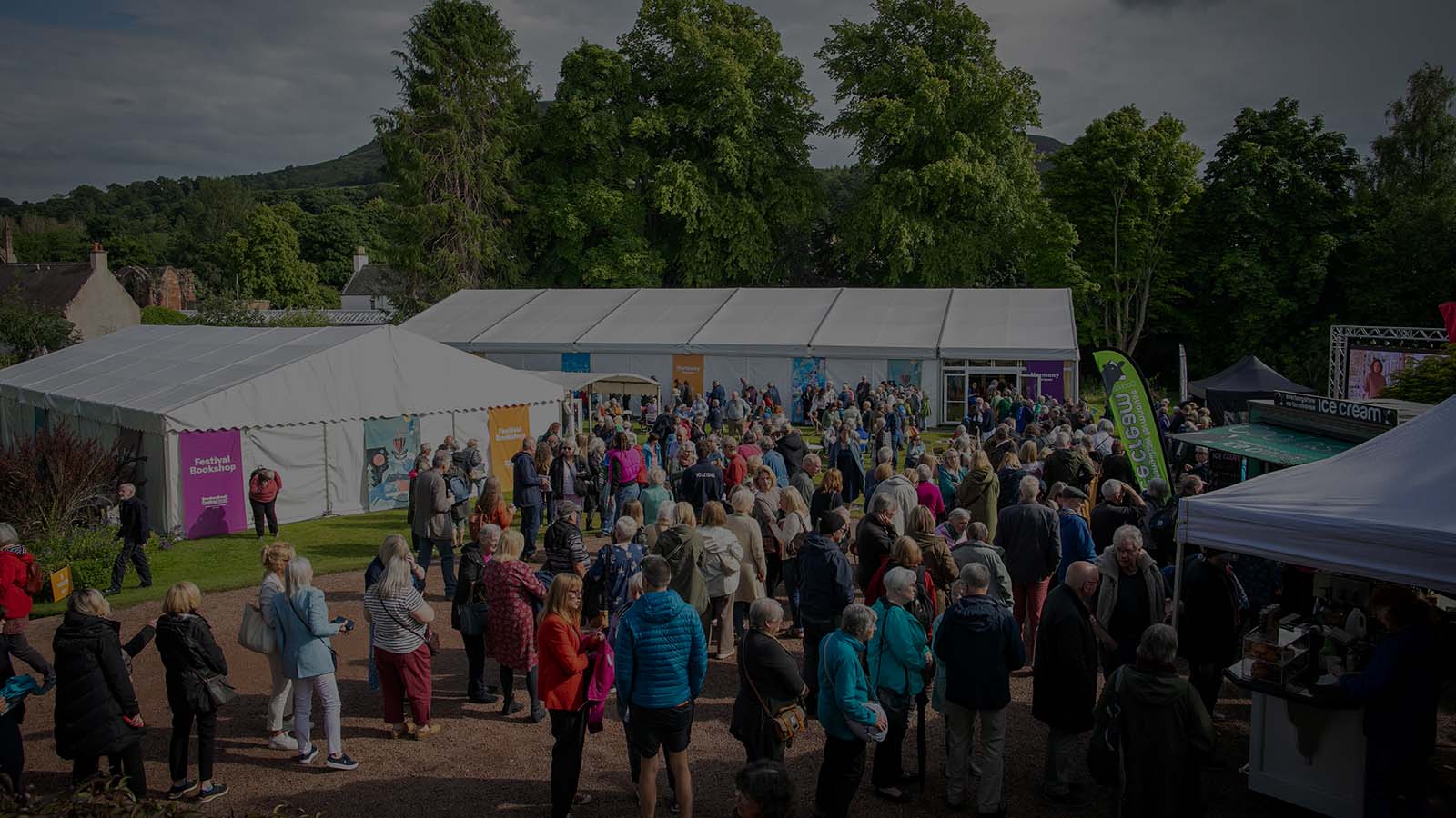 A large crowd gathers outdoors near white tents for a festival, surrounded by greenery and various booths, creating a vibrant atmosphere.