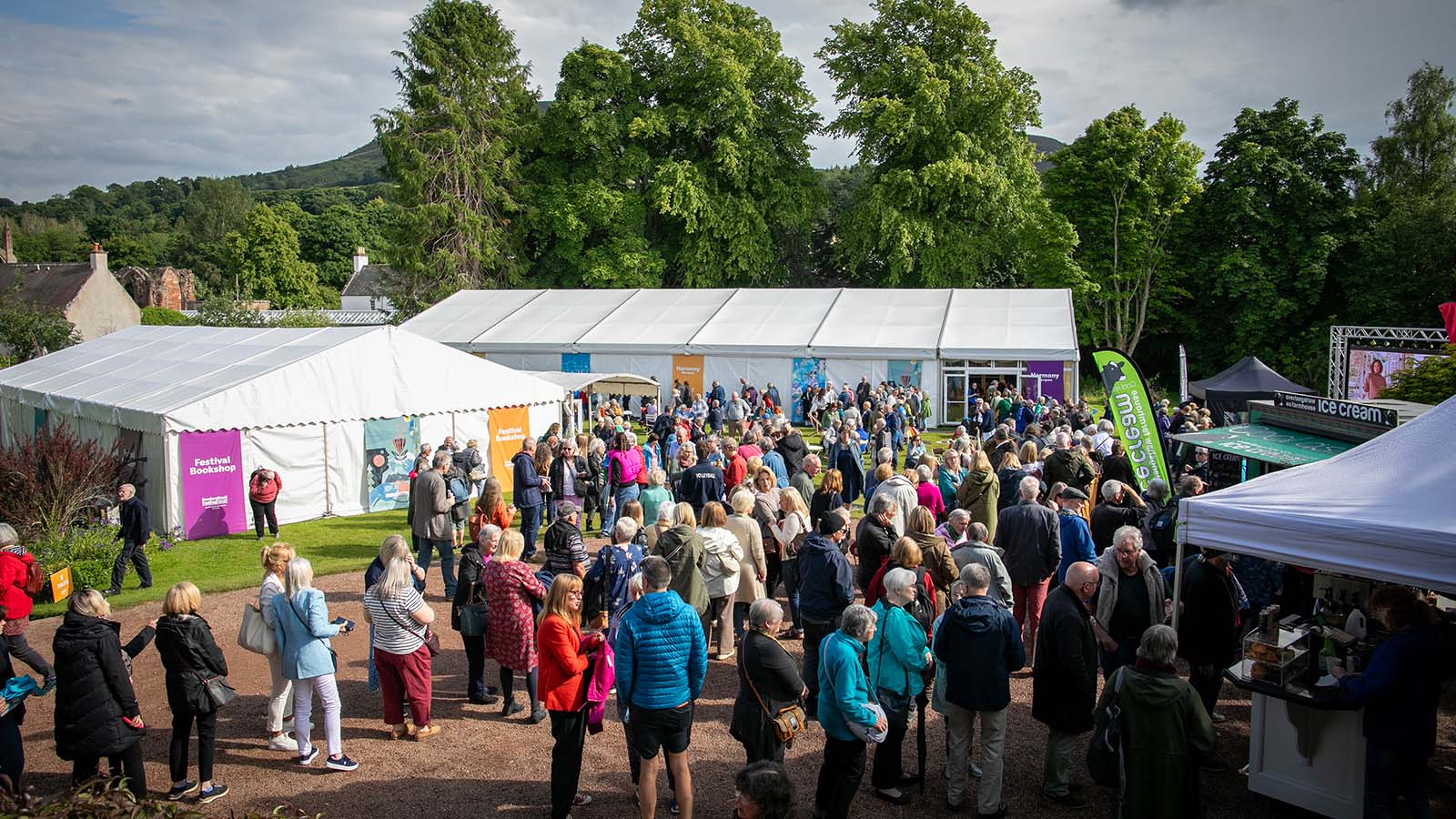 A large crowd gathers outdoors near white tents for a festival, surrounded by greenery and various booths, creating a vibrant atmosphere.