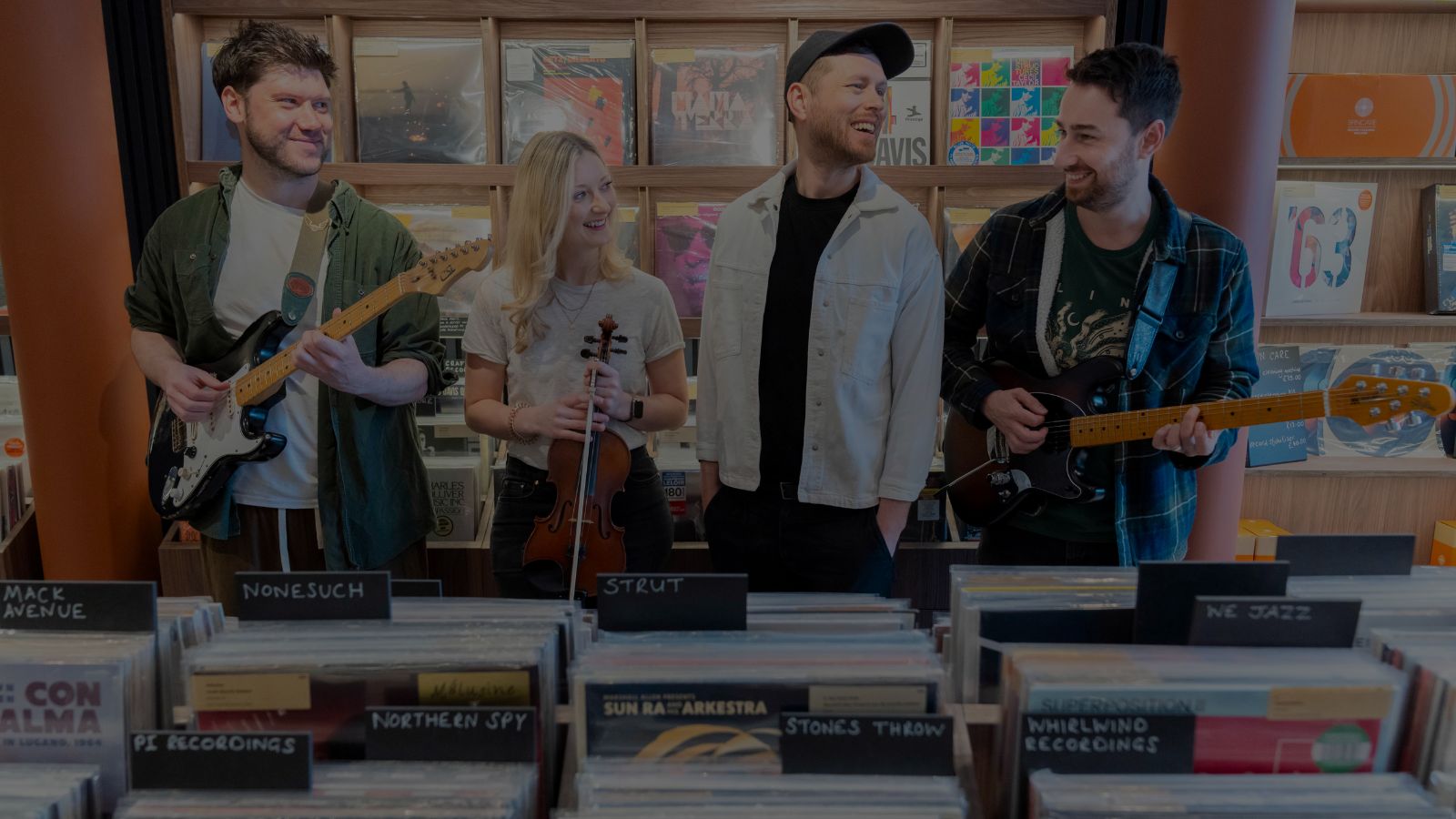A group of musicians stands in a record store, holding instruments with vinyl records displayed prominently in the foreground.