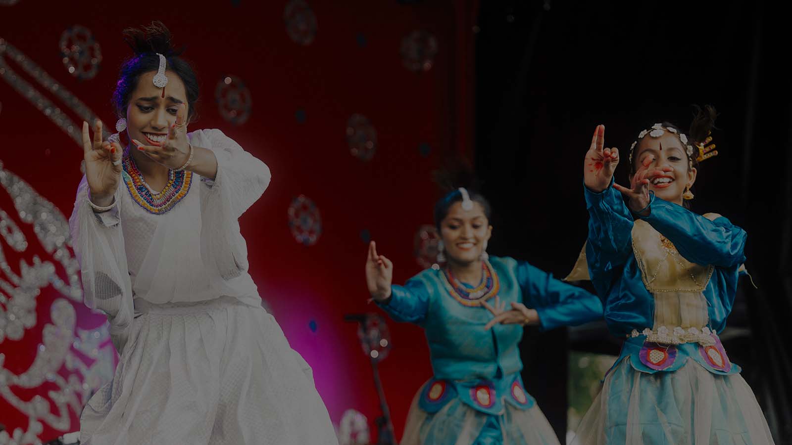 Three dancers in traditional attire perform a lively dance on stage, set against a vibrant red backdrop.