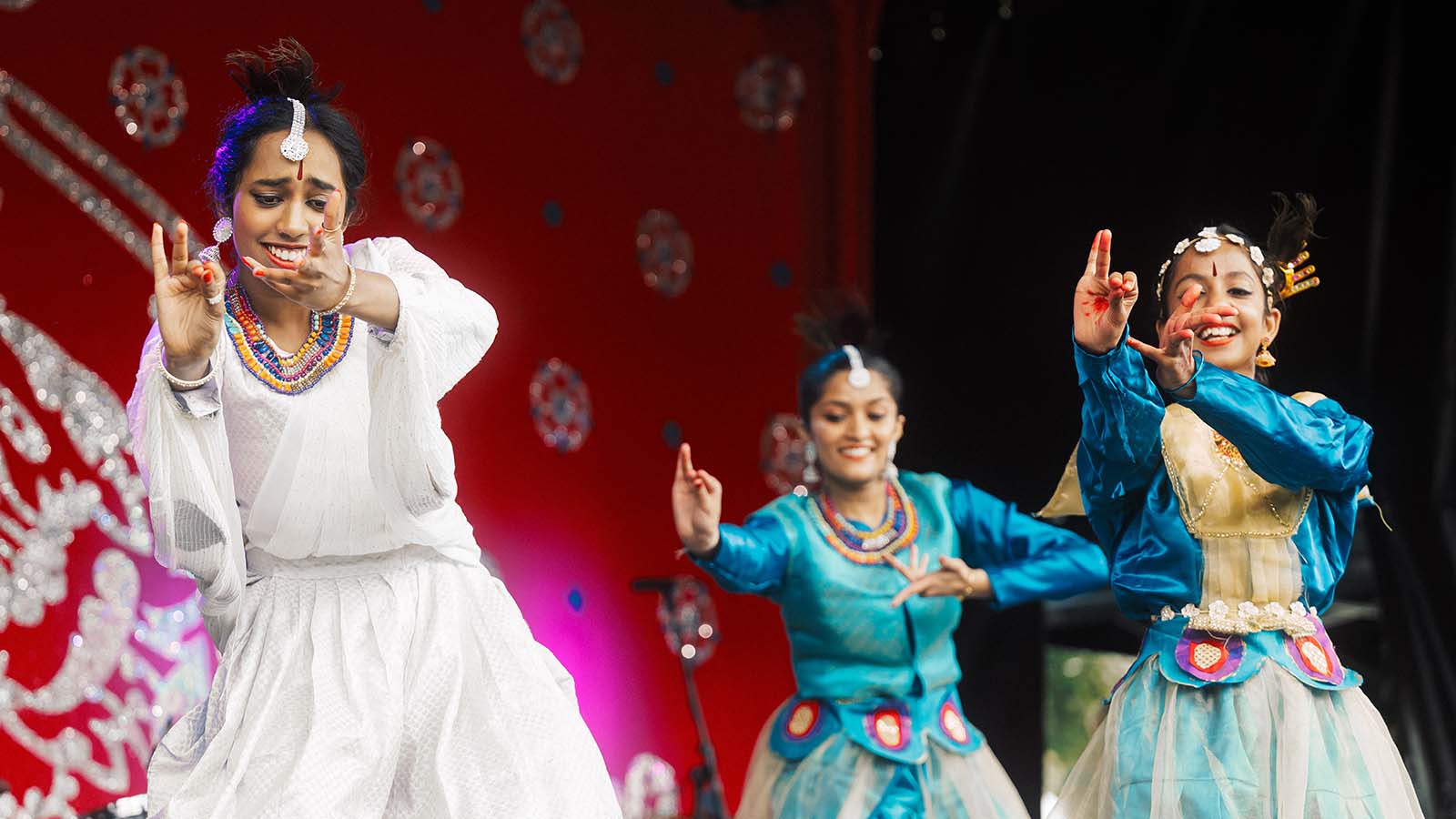 Three dancers in traditional attire perform a lively dance on stage, set against a vibrant red backdrop.