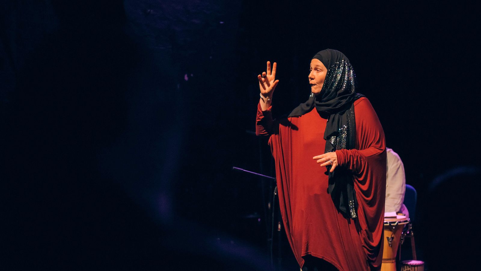 A woman in a flowing red dress and black hijab gestures passionately while performing on stage, with drums visible behind her.