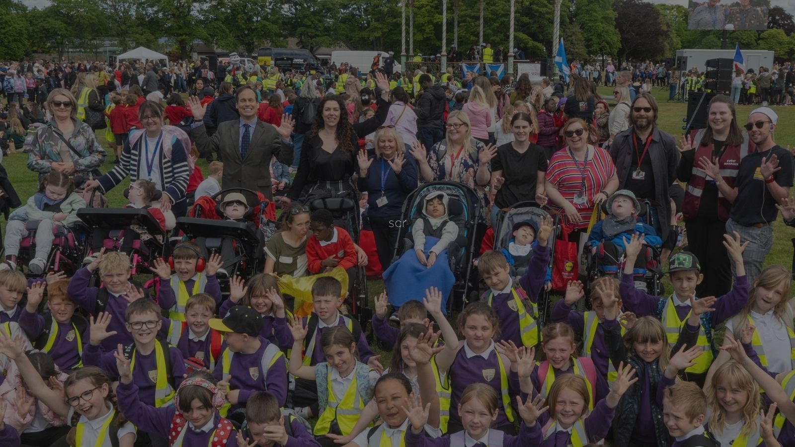 A lively outdoor gathering featuring children in bright vests and adults, all smiling and waving in front of a decorative bandstand.