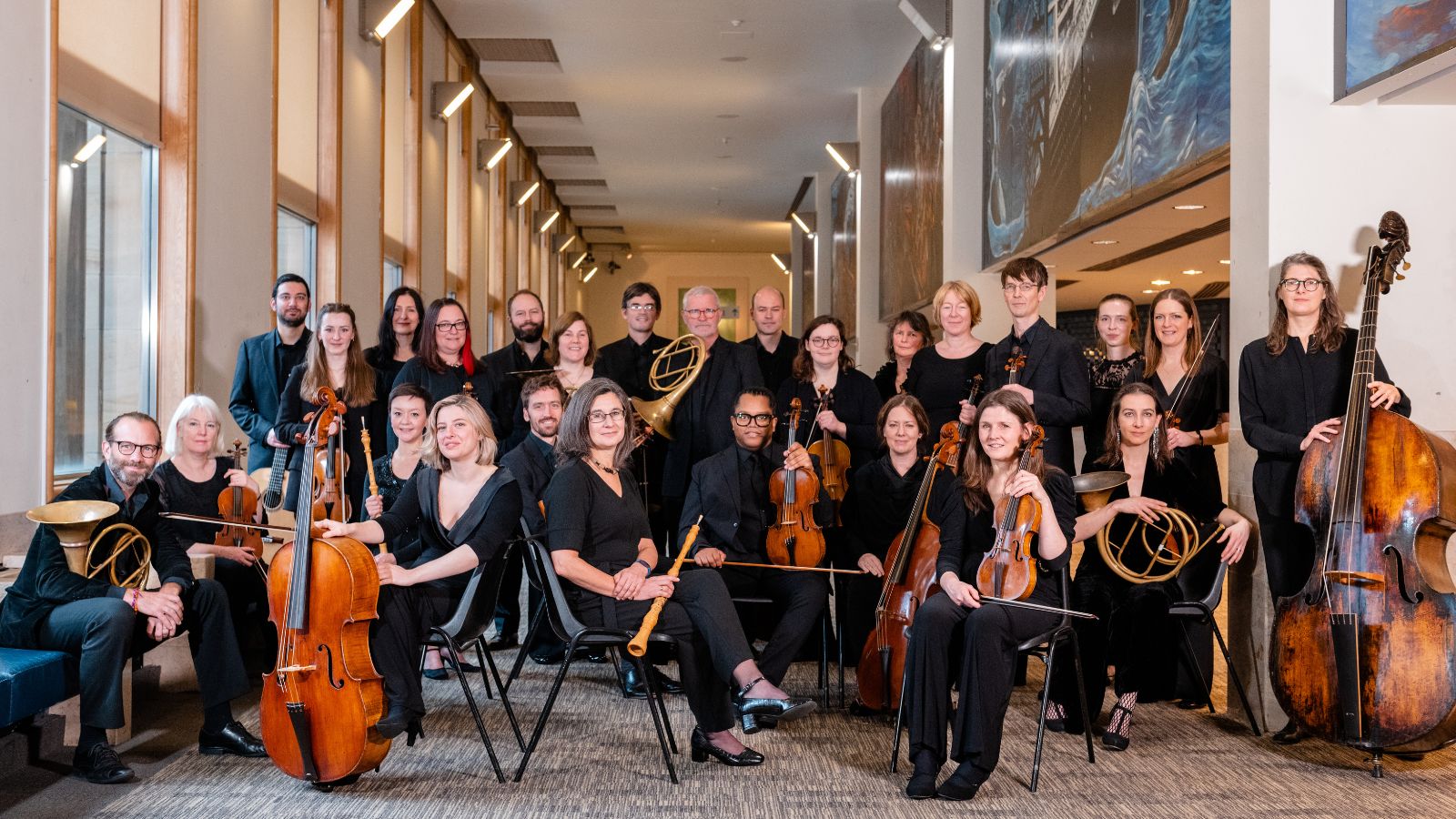 A diverse orchestra posed indoors, dressed in black, with various instruments including strings and brass, against a bright, modern backdrop.