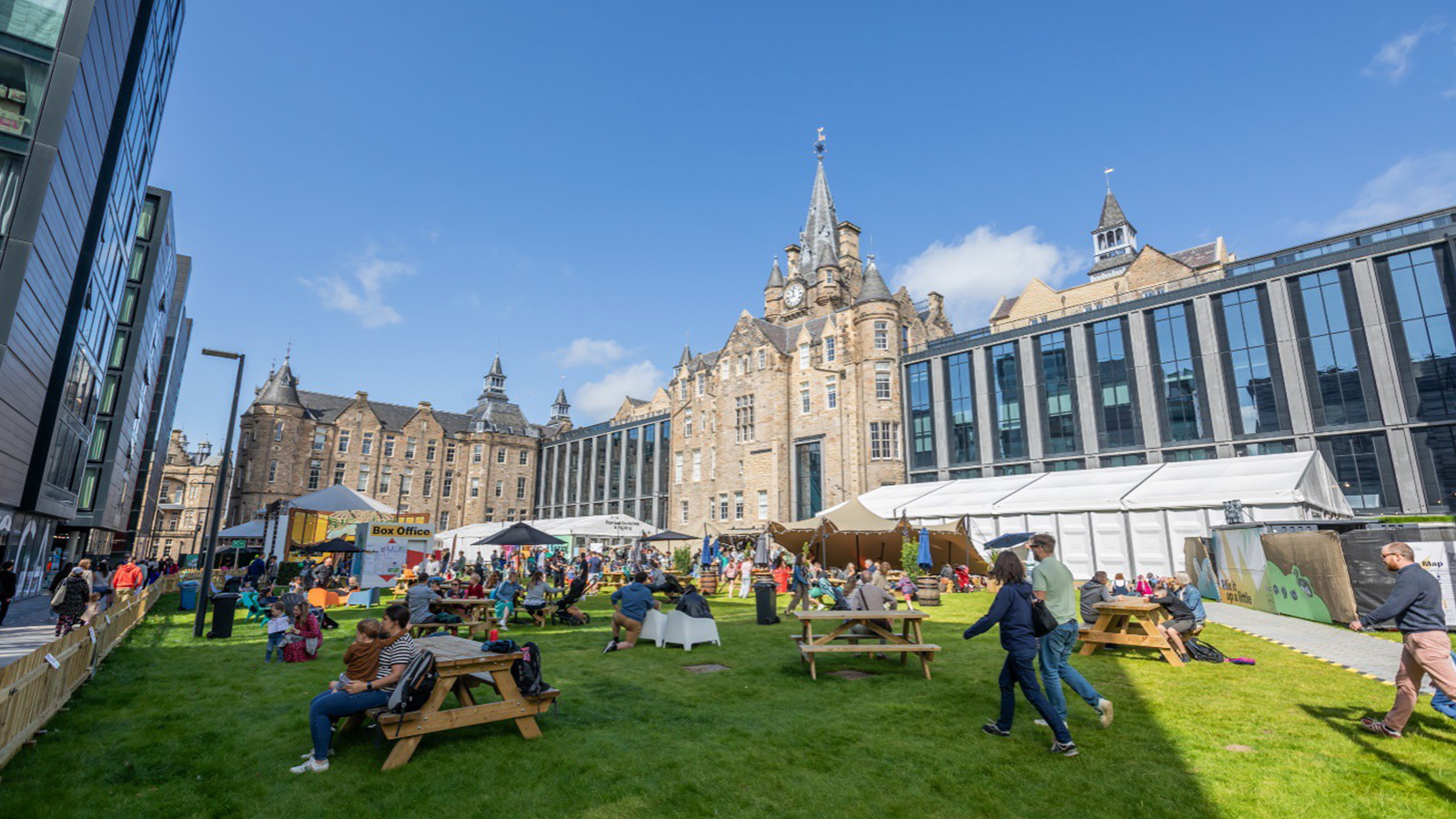 A bustling outdoor market scene in a city park, surrounded by historic and modern buildings, with people enjoying food and activities.