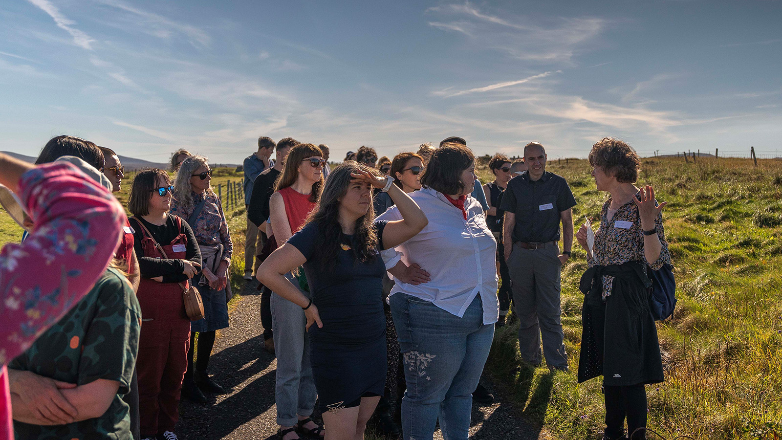 A diverse group of people gathered outdoors, engaged in conversation against a backdrop of green fields and a clear blue sky.